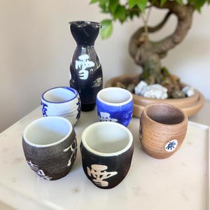 Puede incluir: Un juego de sake japonés con una botella de cerámica negra y blanca y seis tazas a juego. Las tazas son de varios colores, incluyendo blanco, azul y marrón, con caracteres japoneses. El juego está diseñado para servir sake.