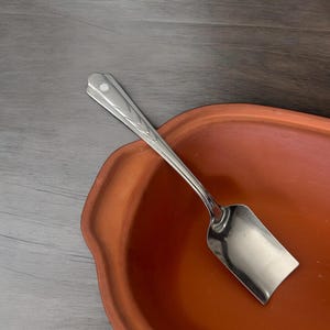 Può includere: Un cucchiaio a paletta in acciaio inossidabile con manico decorativo è appoggiato su un piatto color terracotta. Il cucchiaio ha una paletta rettangolare e un manico con un motivo a spina di pesce. Il piatto ha una forma arrotondata e irregolare.