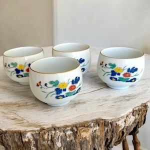 Könnte beinhalten: Vier weiße Keramik-Teetassen mit einem Blumendesign mit blauen, gelben und roten Blumen. Jede Tasse hat einen goldenen Rand und steht auf einer hellen Holzoberfläche. Die Tassen sind in einer Gruppe angeordnet.