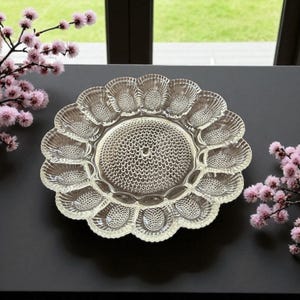 May include: Clear glass serving platter with a scalloped edge and a central textured design. The platter has 12 individual sections around the edge. The platter is on a dark surface with pink flowers in the background.