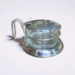 May include: A clear glass serving dish with a polished silver-tone metal frame and a matching hinged lid. The container features a curved metal handle attached to the side, allowing for easy lifting of the lid and transport of the glass bowl.