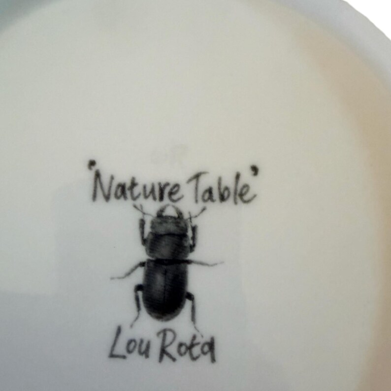 Pu&ograve; includere: Una superficie in ceramica bianca con un'illustrazione stampata in nero di un cervo volante centrata tra il testo Nature Table sopra e il nome Lou Rota sotto. Il design &egrave; reso in uno stile dettagliato e realistico su uno sfondo chiaro.