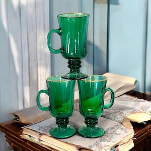 Può includere: Tre tazze di vetro verde con bordi dorati sono impilate. Le tazze hanno manici e basi decorative. Lo sfondo include un muro azzurro e libri aperti. Le tazze sono alte circa 15 cm.
