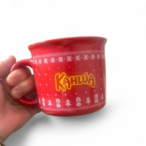 May include: Red ceramic Kahlúa mug with a handle. The mug features the Kahlúa logo in yellow, a white snowflake pattern, and Christmas tree designs. The mug is held by a hand.