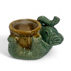May include: A ceramic elephant-shaped planter in shades of green and brown. The elephant is depicted in a seated position, with its trunk curled upwards. The planter has a basket-like design and is suitable for small plants or decorative items.