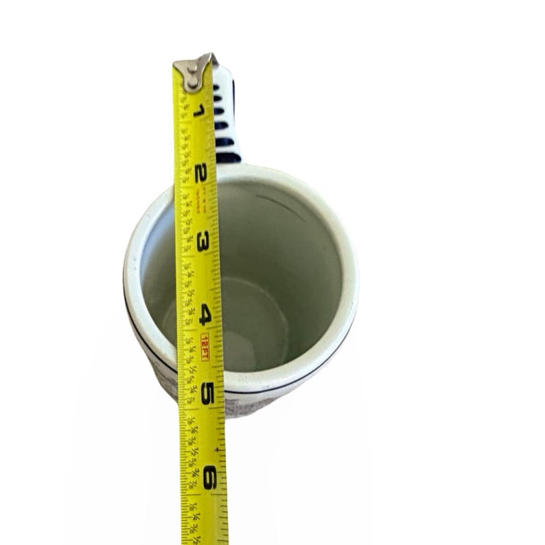 May include: A white ceramic mug with a black rim and handle, measuring approximately 5 inches tall. The mug has a blue and white striped handle and a black design around the base. A yellow measuring tape is placed next to the mug.