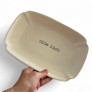 May include: Cream-colored rectangular serving platter with a beaded edge and the words "side kick" printed in black. The platter has a slightly scalloped edge and is held by a hand.