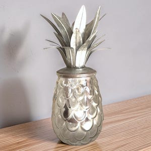 May include: A decorative pineapple-shaped container with a reflective, silver-toned glass body featuring a textured, scalloped pattern. The lid is topped with a metallic, silver-coloured crown of pointed, upright leaves made of thin metal.