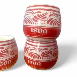 May include: Three white and red tequila shot glasses stacked on top of each other. The shot glasses have a red band with the text "1800 TEQUILA" and a decorative floral design. The shot glasses are barrel-shaped.
