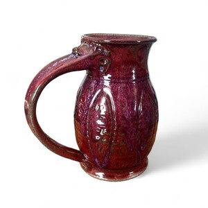 May include: A handcrafted, burgundy-colored ceramic mug with a large handle and a detailed, textured design. The mug has a glossy finish and a unique, sculpted detail where the handle meets the body. The base is a darker shade of red.