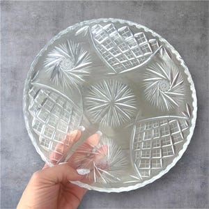 May include: A clear, round glass serving plate with intricate etched patterns. The design includes starbursts, diamond shapes, and spiral details. The plate has a scalloped edge and is held by a hand.