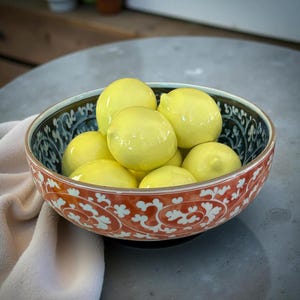 May include: A decorative ceramic bowl filled with yellow lemons. The bowl has a red and white floral pattern on the outside and a blue and white design on the rim. The bowl sits on a gray surface.