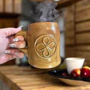 May include: A brown ceramic mug with a handle, embossed with a floral design, and filled with steaming liquid. The mug is held in a hand with long nails. The mug is on a wooden surface.