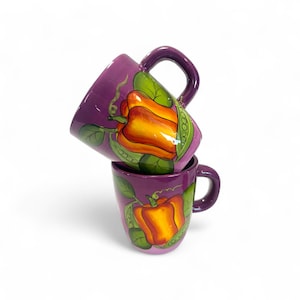 May include: Two purple ceramic mugs stacked, each with a handle and a painted design. The design features a yellow and orange bell pepper with green leaves and peas. The mugs are on a white background.