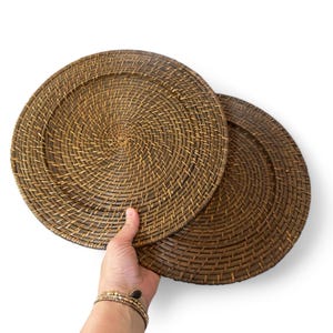 May include: Two round, woven placemats in a warm brown hue. The placemats are made of natural material and feature a spiral weave pattern. The placemats are held by a hand with a bracelet. The background is white.
