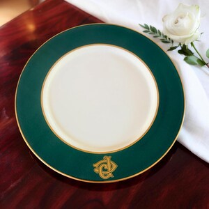 May include: A round, white plate with a dark green border and gold trim. The plate features a gold emblem at the bottom. A white rose and greenery are in the background. The plate is on a dark wood surface.