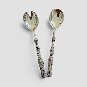 Pode incluir: Um par de talheres de salada prateados. Um tem um design de colher com fenda, e o outro tem um design de colher sólido. Ambos os talheres têm cabos ornamentados e texturizados com aparência de borla. As colheres são reflexivas.