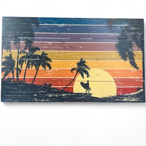 May include: Wooden wall art featuring a sunset scene with a surfer, palm trees, and a large sun. The artwork has a distressed, vintage look with horizontal stripes of color in shades of blue, orange, and yellow.