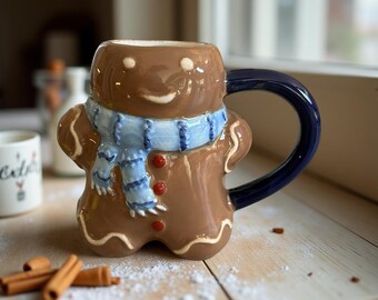 以下が含まれることがあります： 青いスカーフと濃い青色のハンドルが付いた、茶色のセラミックジンジャーブレッドマンマグ。マグには笑顔の顔と赤いボタンのディテールがあります。手前にはシナモンスティックがあります。