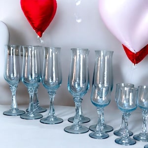 May include: A collection of clear, stemmed glassware with a light blue tint. The glasses vary in size and shape, including champagne flutes and smaller wine glasses. Red and pink heart-shaped balloons are in the background.