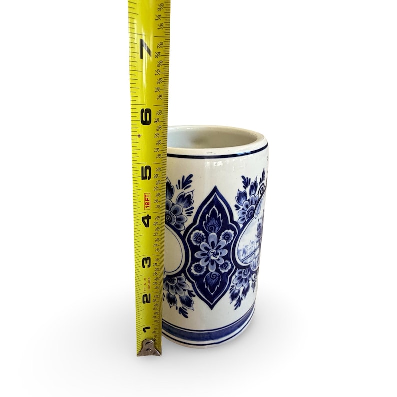 May include: A white and blue ceramic mug with a floral design and a landscape scene. The mug is approximately 5 inches tall. The mug has a cylindrical shape and a blue rim.