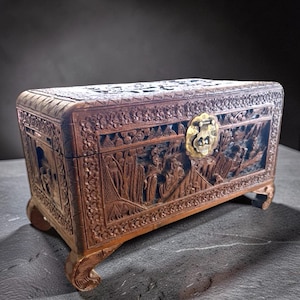 May include: A rectangular wooden chest featuring intricate relief carvings of figures and landscapes on all visible sides. The lid is domed and the front includes a central brass latch mechanism. The chest is supported by four curved feet.