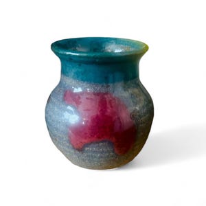 May include: A small, handcrafted ceramic vase with a rounded body and a wide opening. The top is teal, transitioning to a gray body with a vibrant red splash. The vase is suitable for displaying small flowers or as a decorative piece.
