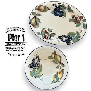 May include: Two pieces of Pier 1 earthenware. An oval platter and a round bowl feature a cream-coloured background with a fruit design. The fruit includes pears, plums, grapes, and cherries. The rims have a dark blue border. The text "Pier 1" is visible.