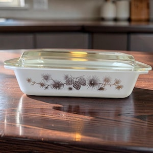 May include: A rectangular white glass baking dish with a clear glass lid. The exterior of the white dish features a brown printed pattern of pine cones and pine needles centered along the side. The dish has integrated handles on both ends.