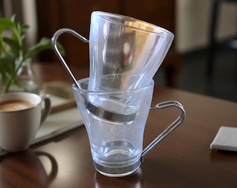 Op de afbeelding: Twee doorzichtige glazen koffiekopjes die op elkaar zijn gestapeld. Elk kopje heeft een zilveren handvat en een zilveren band in het midden. De kopjes staan op een donkerbruin houten oppervlak.
