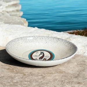 May include: A white ceramic bowl with a speckled rim and a central design. The design features a figure, mountains, and a pot within a brown and blue circle. The bowl is set on a stone surface with a blue water background.