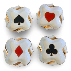 Puede incluir: Cuatro platos blancos en forma de trébol con ribete dorado, cada uno con un símbolo de palo de cartas diferente: picas, corazones, diamantes y tréboles. Los palos son negros y rojos.