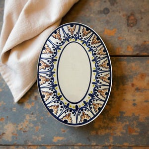 May include: Oval ceramic serving platter with a cream-colored center and a decorative border. The border features a repeating pattern of brown, blue, and yellow floral motifs. The platter is set on a distressed wooden surface, with a beige linen napkin in the background.
