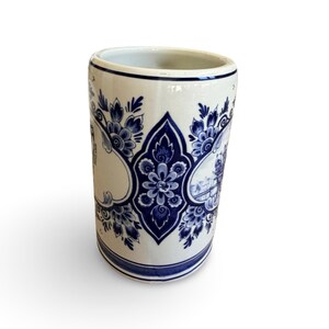 May include: A white ceramic cylindrical vase with blue floral and architectural designs. The vase features a repeating pattern of flowers and a detailed illustration of a building. The rim and base are accented with blue lines.
