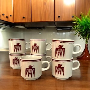 May include: A set of six white ceramic mugs with a dark red bird design. The mugs have a handle and a dark red band around the rim. The bird design is a stylized image of a bird with a geometric pattern.