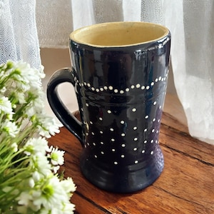 May include: A dark blue ceramic mug with white polka dots and a handle. The interior is a light tan color. The mug is on a wooden surface, with white flowers and a sheer white curtain in the background.