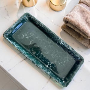 May include: A rectangular, dark green tray with a marbled pattern and gold trim. The tray is on a white marble surface, with a stack of folded towels and gold-colored containers in the background.