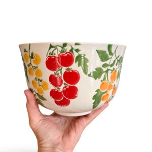 Puede incluir: Un cuenco grande de cerámica color blanco roto con un patrón pintado a mano de tomates cherry rojos y amarillos sobre vides verdes. El diseño envuelve el exterior del cuenco, mostrando racimos de frutos redondos y hojas verdes detalladas.