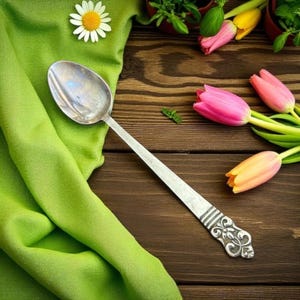 May include: A silver spoon with an ornate handle rests on a wooden surface next to a green cloth, a daisy, and colorful tulips. The spoon's bowl reflects light, and the handle features a detailed floral design. The scene evokes a sense of rustic elegance.