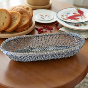 Puede incluir: Una cesta de pan de malla de alambre plateada con forma ovalada. La cesta está sobre una mesa de madera, con rebanadas de pan y platos decorativos en el fondo. La cesta está vacía.