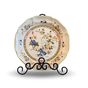 May include: A decorative, octagonal porcelain plate with a floral design, displayed on a black metal stand. The plate features a cream-colored background with a border of flowers and a central motif of blue, yellow, and orange blossoms.