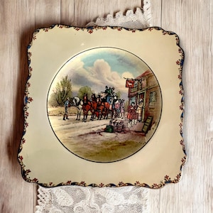 Könnte beinhalten: Quadratischer cremefarbener Teller mit einer detaillierten Illustration einer Pferdekutsche vor einem Gebäude. Der Teller hat einen gewellten Rand mit roten und blauen Blumenakzenten und einen blauen Rand.