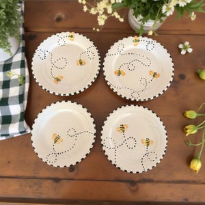 May include: Four white ceramic plates with a black checkered rim. Each plate features a whimsical design of a bee and dotted flight path in yellow and black. The plates are arranged on a wooden surface, suggesting a kitchen or dining setting.