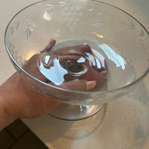 Puede incluir: Un cuenco de vidrio transparente con diseños grabados de uvas y hojas alrededor del borde. El cuenco tiene una base ancha y poco profunda y una base resistente. El vidrio refleja la luz, creando reflejos y sombras.