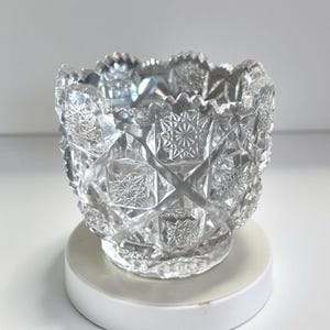 May include: A clear, cut-glass bowl with a detailed geometric pattern. The bowl has a scalloped rim and sits on a small, round base. The intricate design features starburst and diamond shapes, creating a sparkling effect. This decorative bowl is ideal for display.