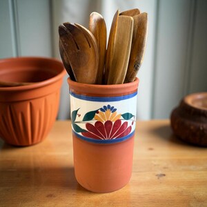 May include: A terracotta utensil holder with a floral design in red, yellow, and blue. The holder contains several wooden kitchen utensils. The design includes a blue and white band around the middle. The holder is on a wooden surface.