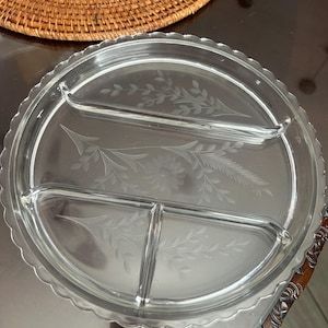 May include: Clear glass serving tray with three compartments and a scalloped edge. The tray features etched floral and leaf designs. A woven placemat is visible in the background.