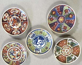 Può includere: Cinque piatti decorativi in ceramica con intricati disegni floreali e geometrici. I piatti presentano una varietà di colori, tra cui rosso, blu, verde e oro, con motivi dettagliati e un bordo color crema. I piatti sono disposti su una superficie grigia.