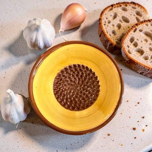 May include: A round ceramic grater plate with a bright yellow interior and a brown rim. The center features a raised, textured brown surface for grating. The plate is surrounded by three whole garlic bulbs and two slices of crusty sourdough bread.