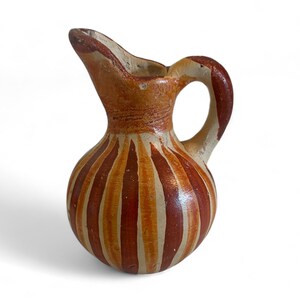 May include: A small, hand-painted ceramic jug with a rounded body and a handle. The jug is decorated with vertical stripes in shades of brown and cream. The spout is slightly chipped, adding to its rustic charm.